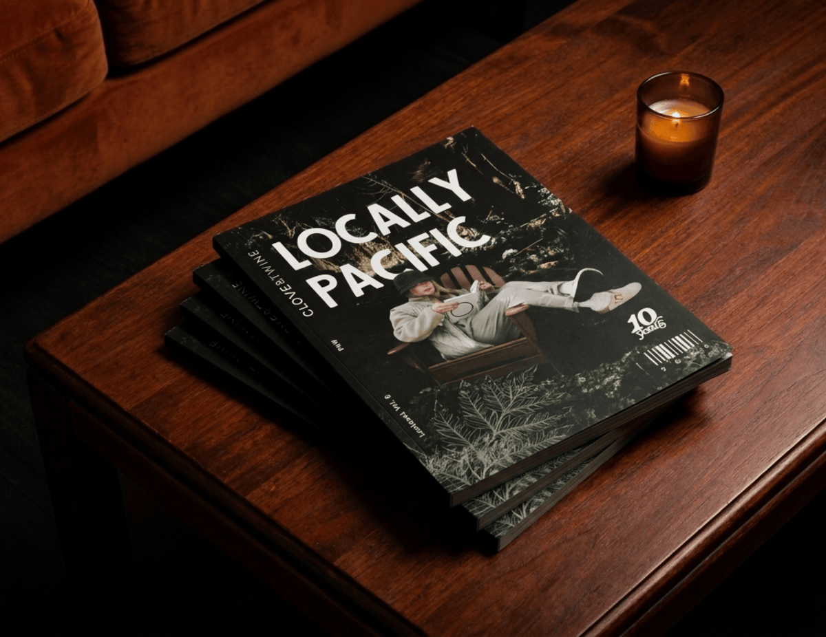 A stack of printed magazines or brochures titled "Locally Pacific" sits on a dark wooden table beside a lit amber candle, with part of a brown upholstered chair visible in the upper left.